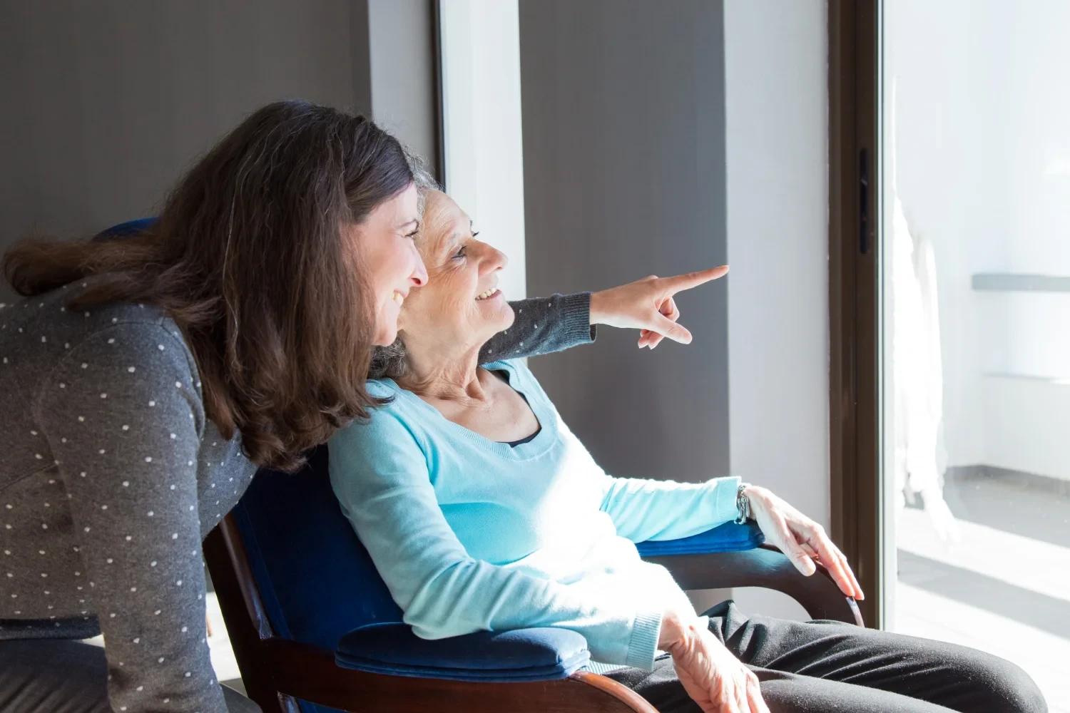 A caregiver and elderly woman enjoying a moment together, looking outside from a comfortable chair.