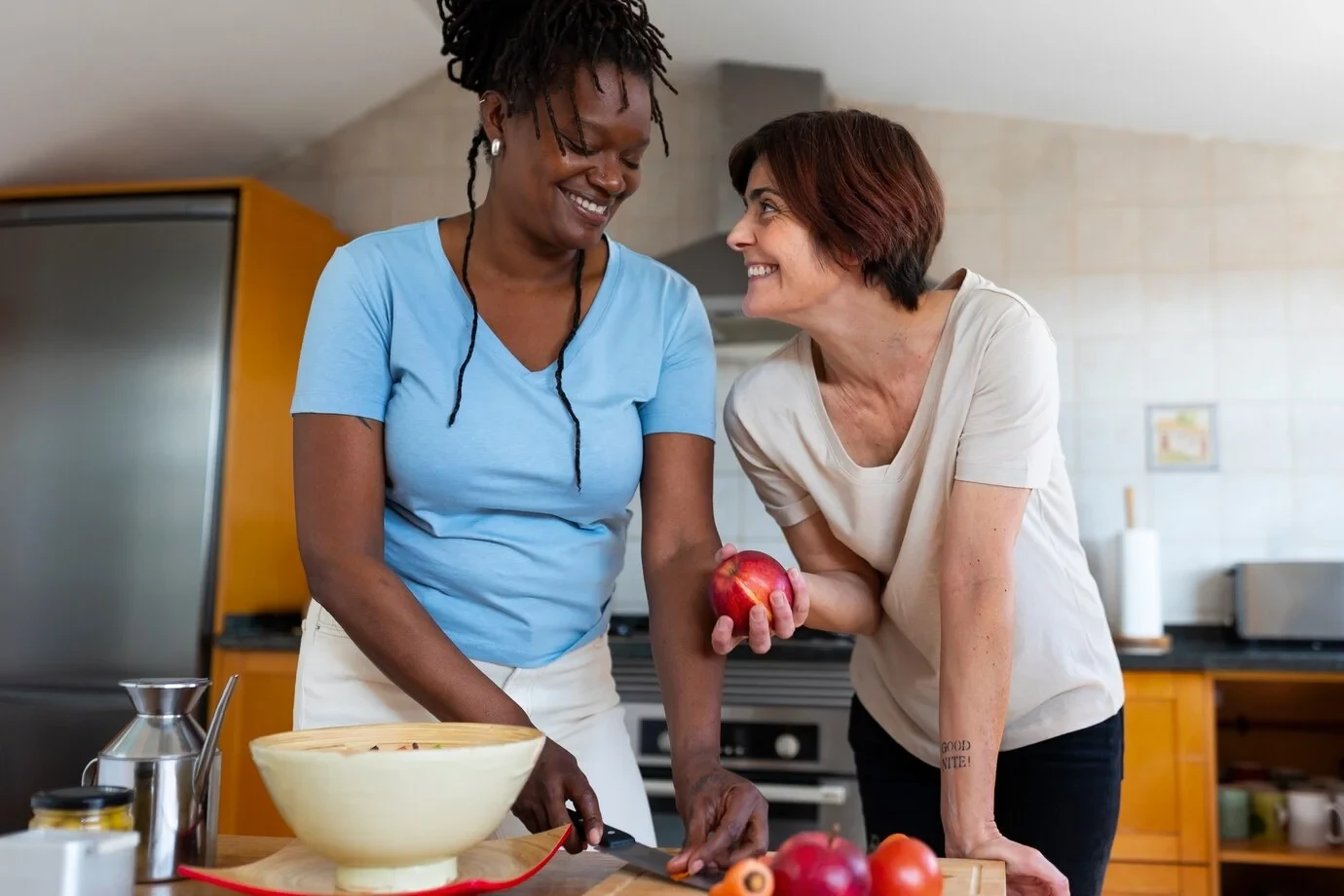 Two women smiling and preparing food together in a kitchen, reflecting companionship and support in assisted living.