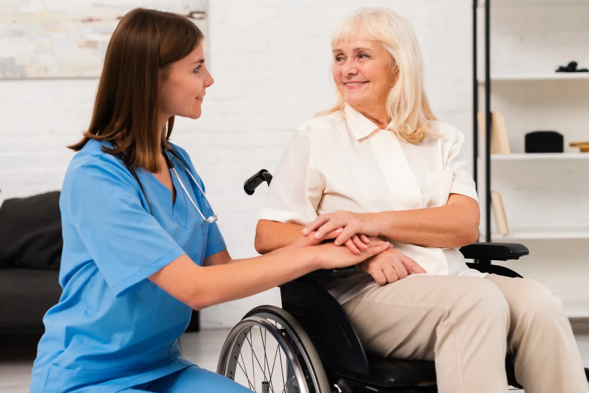 Healthcare worker smiling and holding hands with a woman in a wheelchair, showing warmth and connection in a common area.