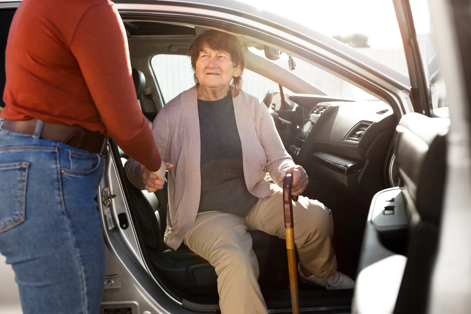 Resident woman assisted by caregiver stepping out of car using cane for transportation service
