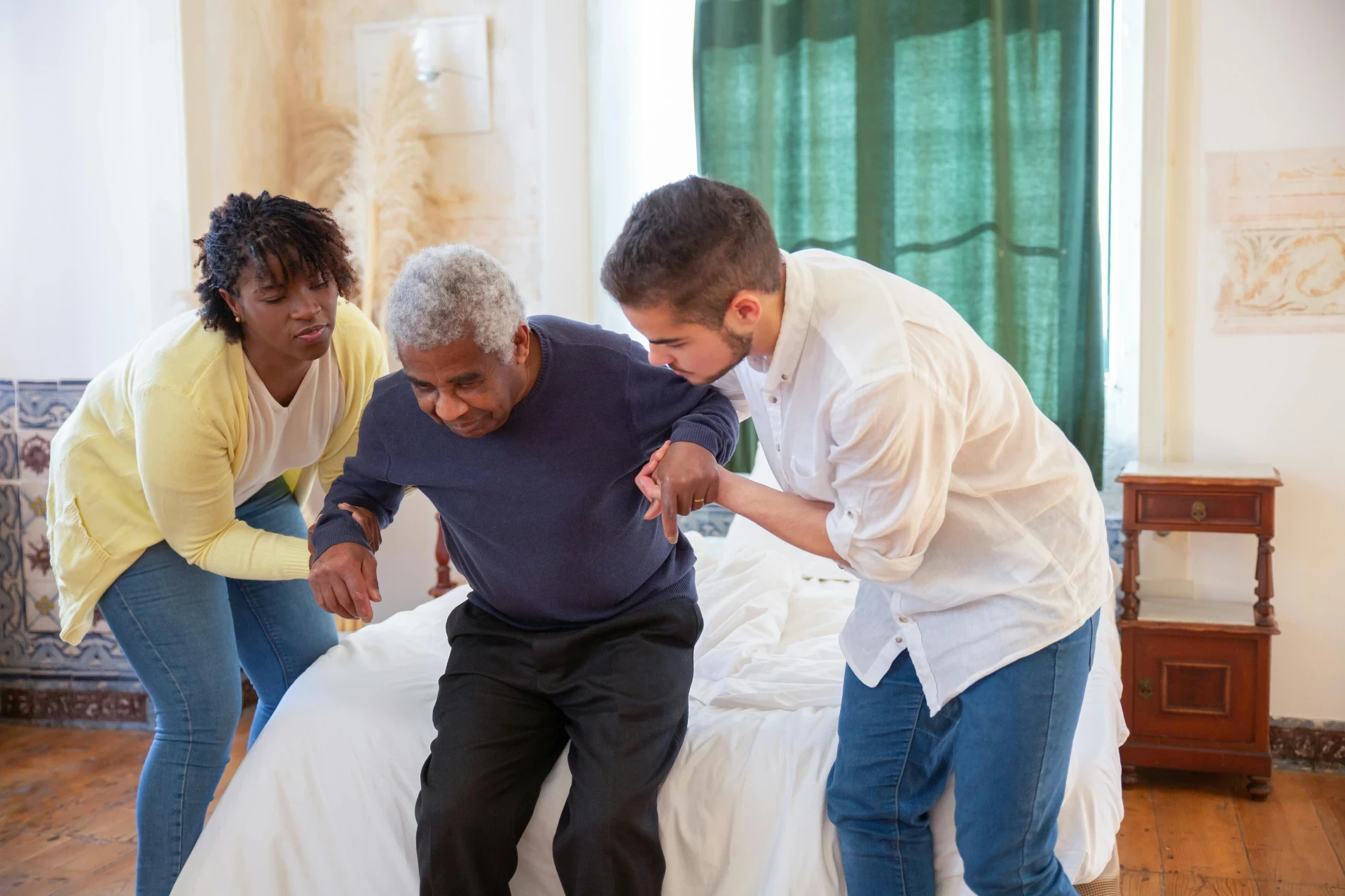 Two caregivers assisting an elderly man in standing up from a bed, providing mobility support in a home setting.
