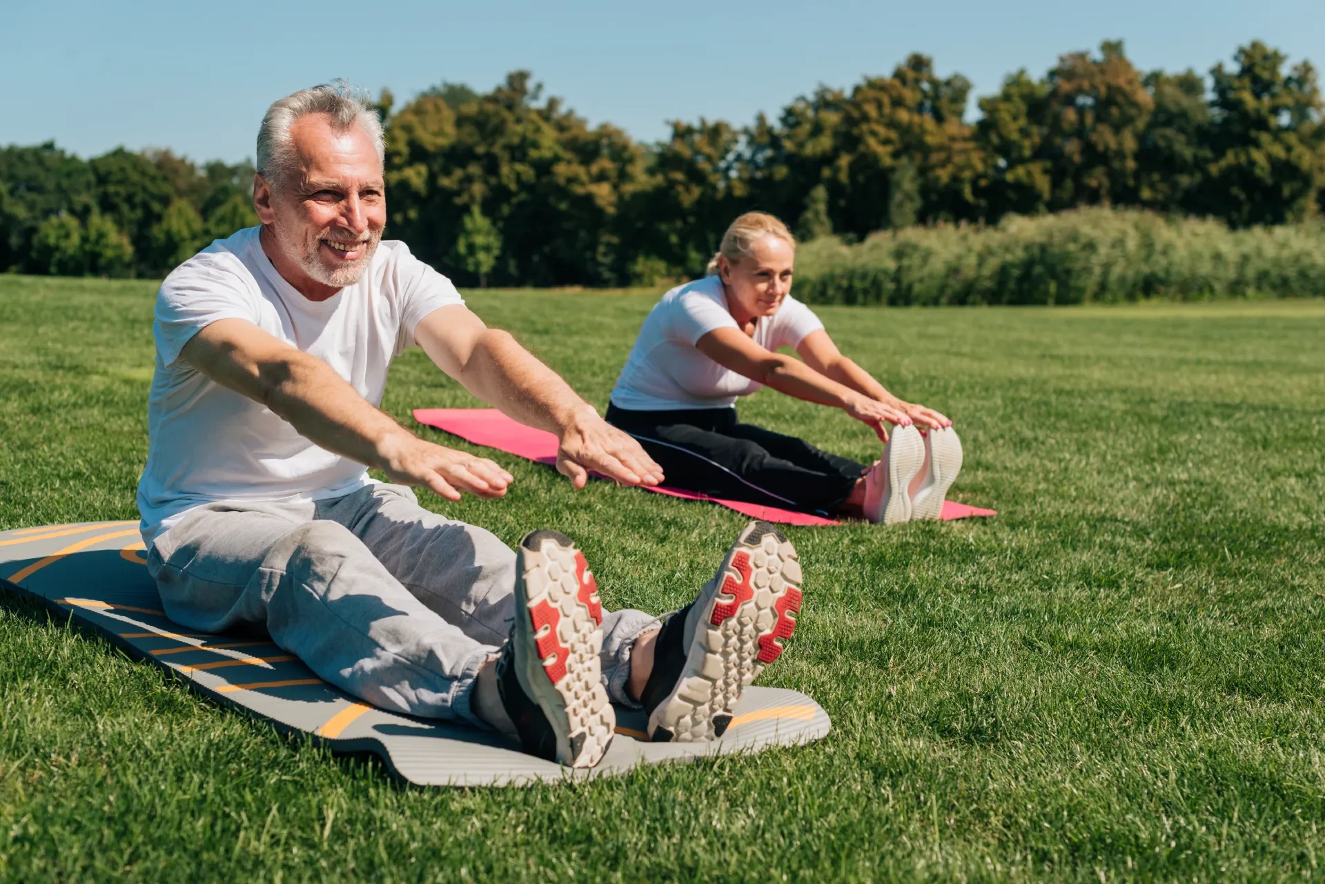 Two older adults stretching on exercise mats in a sunny open field, promoting active living in outdoor spaces.