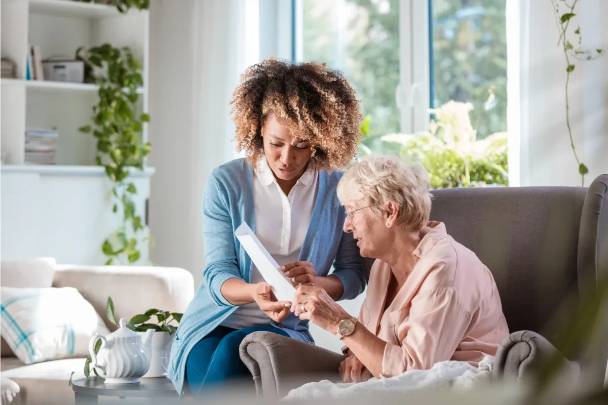 Caregiver sitting with an elderly woman on a couch, reviewing a document together in a well-lit, comfortable living space.